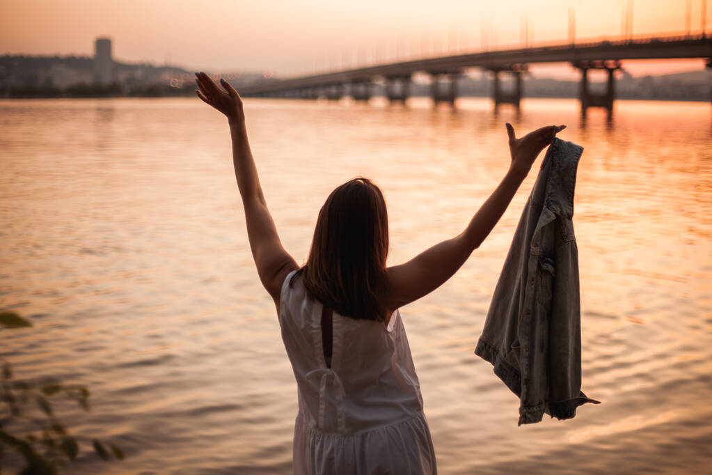 una giovane donna in abito bianco con le mani alzate sta ballando su un tramonto arancione in città e il fiume e il ponte - Foto, immagini