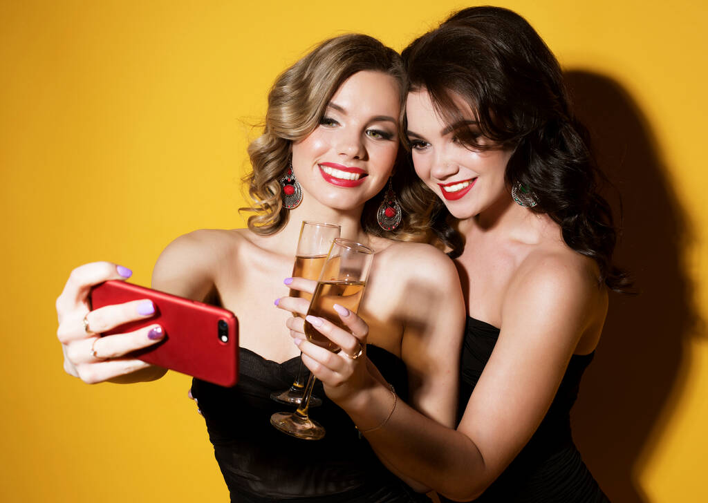 twee chique jonge vrouwen in zwart cocktail jurken drinken champagne en het nemen van een selfie. - Foto, afbeelding