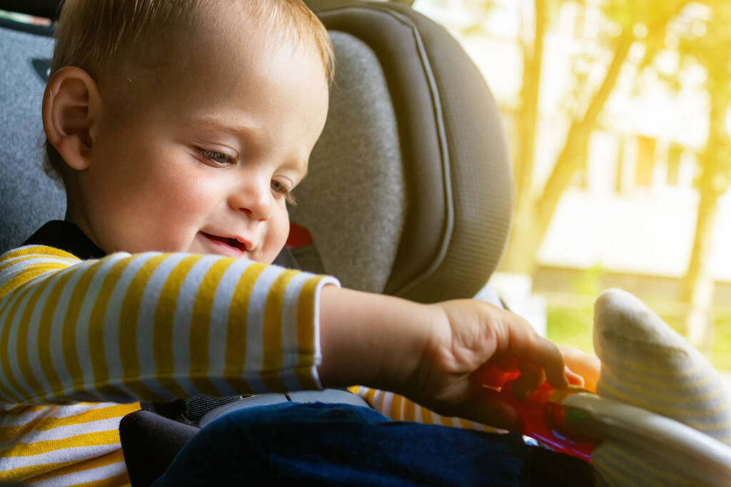 Αγνώριστο καυκάσιο μωρό κάθεται σε κάθισμα αυτοκινήτου, κρατώντας παιχνίδι στα χέρια, παίζοντας και χαμογελώντας. Παιχνίδια για ταξίδια με αυτοκίνητο με παιδιά. Καλοκαιρινές διακοπές.  - Φωτογραφία, εικόνα