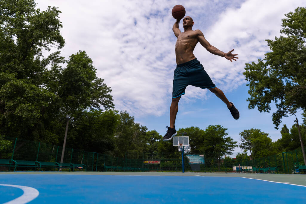 Młody mężczyzna, muskularny afrykański koszykarz grający w koszykówkę na stadionie, boisku sportowym lub na placu zabaw na świeżym powietrzu. Letnie gry sportowe. - Zdjęcie, obraz