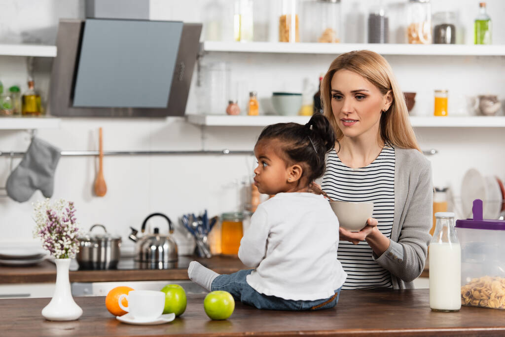 женщина держит миску и смотрит на усыновленного африканского американского ребенка, сидящего на кухонном столе - Фото, изображение