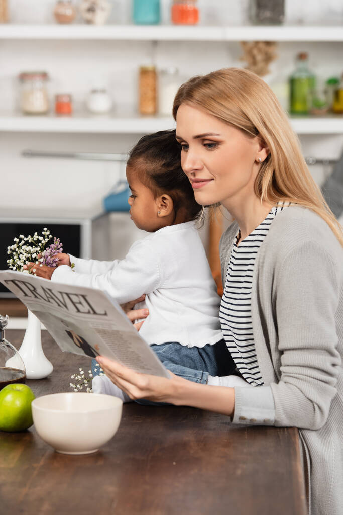 fröhliche Mutter liest Reisezeitung in der Nähe von adoptiertem afrikanisch-amerikanischem Kind in der Küche - Foto, Bild