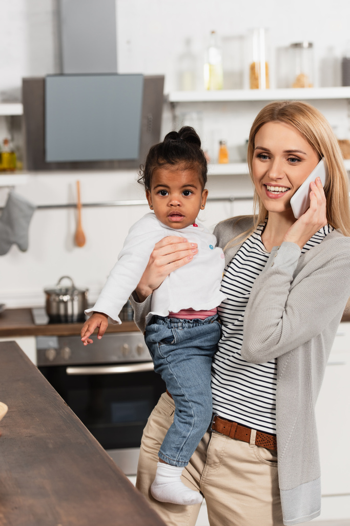 mãe feliz falando no smartphone e segurando adotada criança americana africana na cozinha  - Foto, Imagem