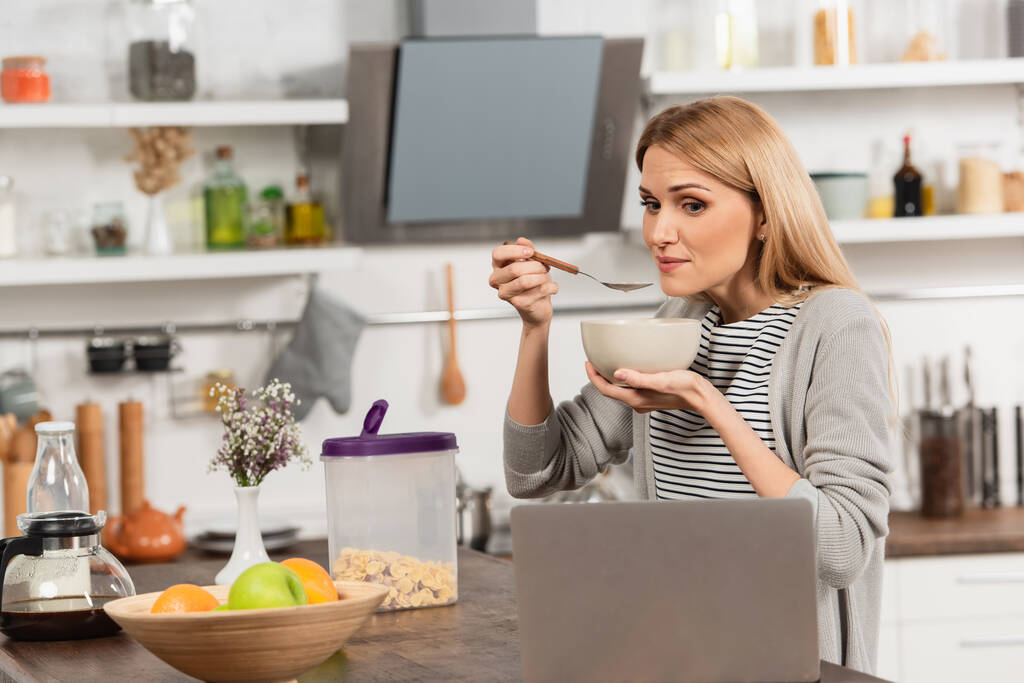 ξανθιά γυναίκα παίρνει πρωινό και βλέποντας ταινία στο laptop στην κουζίνα - Φωτογραφία, εικόνα