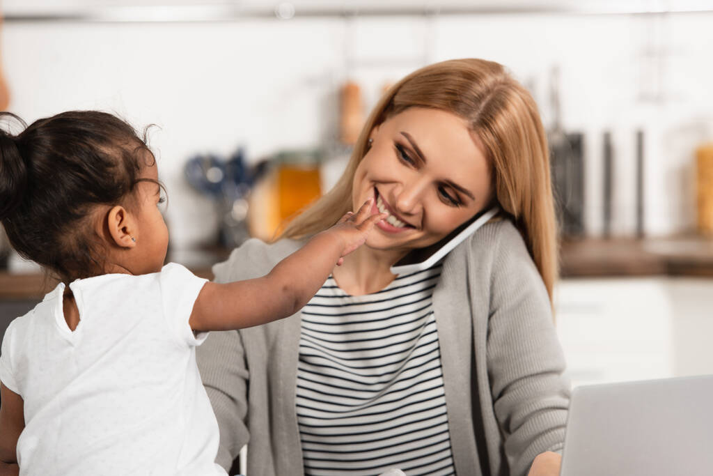 gelukkig moeder praten op smartphone in de buurt van geadopteerd Afrikaans Amerikaans kind met uitgestrekte hand  - Foto, afbeelding