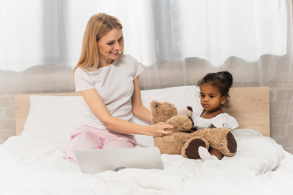 onnellinen äiti tilalla nallekarhu lähellä hyväksyi afrikkalainen amerikkalainen lapsi katsomassa elokuvaa kannettavan makuuhuoneessa  - Valokuva, kuva
