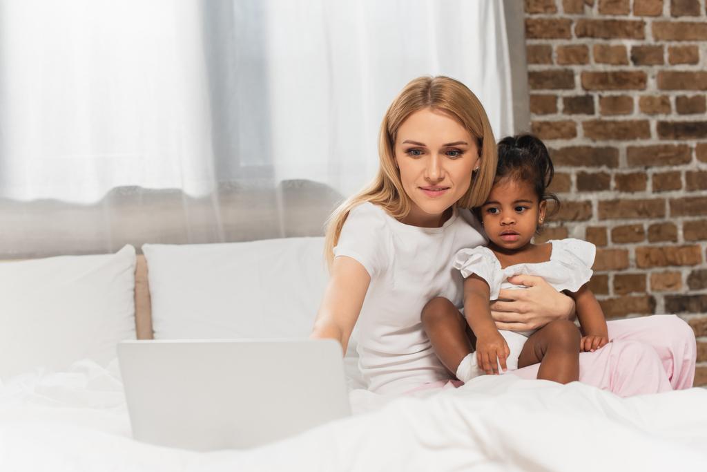wesoła matka i adoptowany afrykański amerykański oglądać film na laptopie w sypialni  - Zdjęcie, obraz