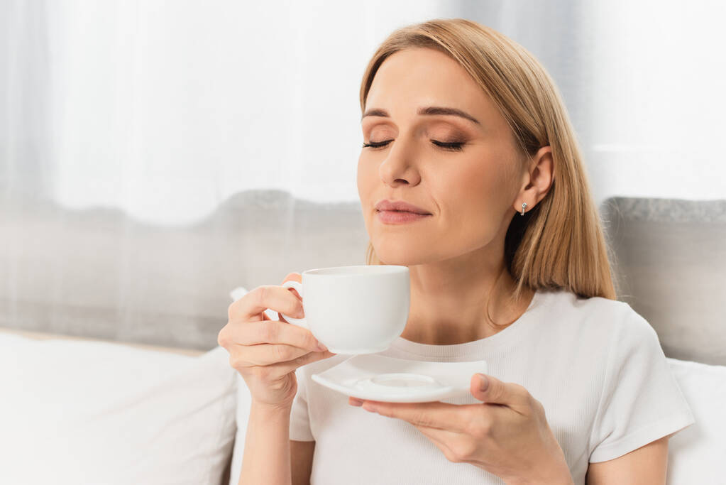 mujer rubia complacida sosteniendo taza de café en el dormitorio  - Foto, imagen