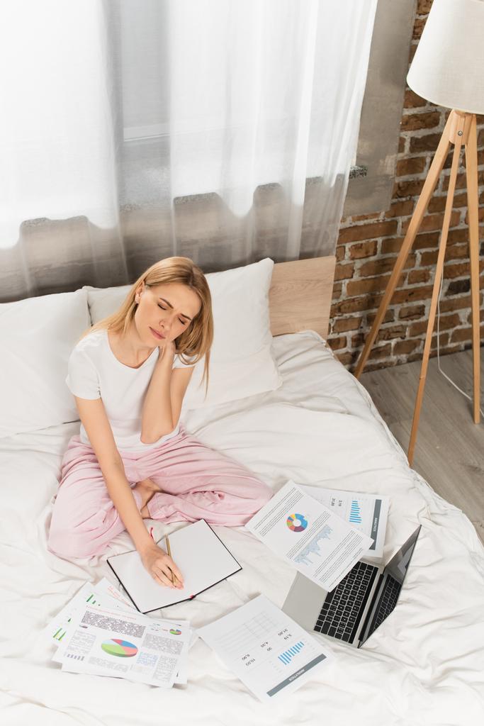 высокий угол обзора усталой женщины, сидящей рядом с графиками и графиками во время использования ноутбука и работы из дома в спальне  - Фото, изображение