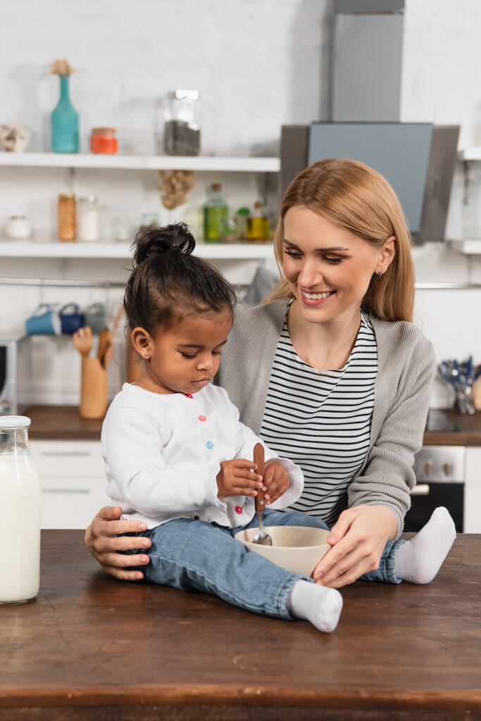 glückliche Mutter blickt auf adoptiertes afrikanisch-amerikanisches Kind mit Löffel auf Küchentisch neben Schüssel sitzend - Foto, Bild