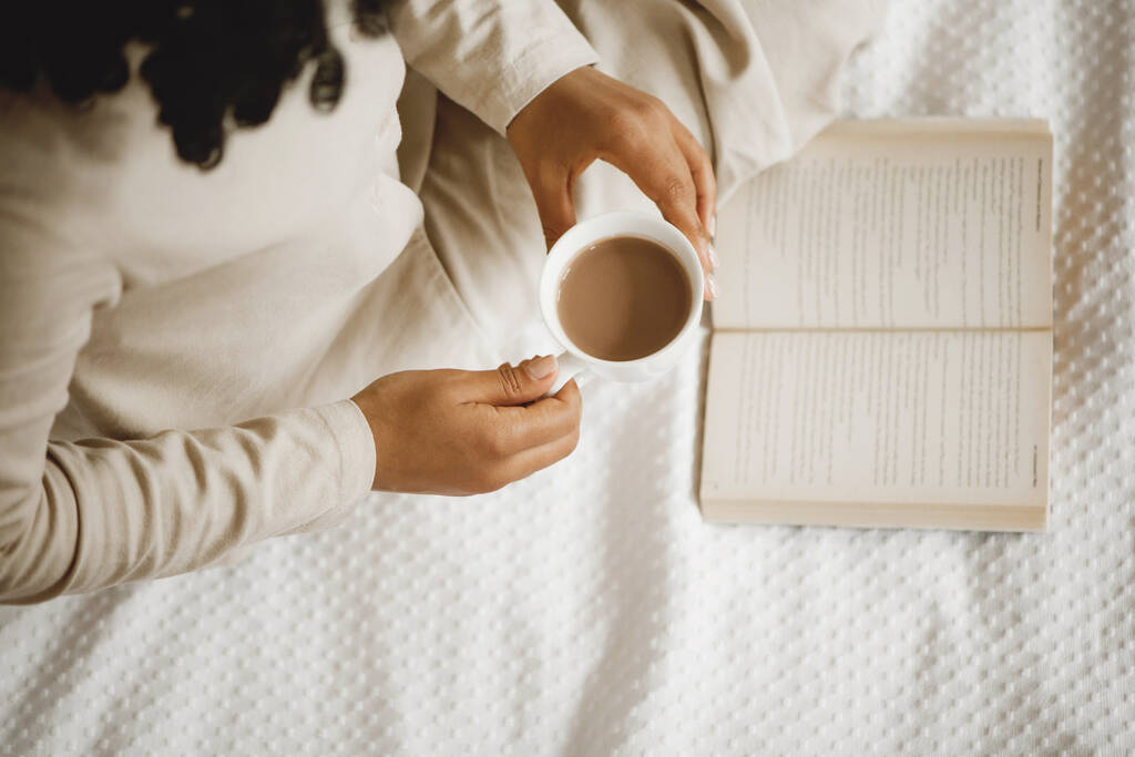 Afroamerikkalainen nainen valkoisella sängyllä juomassa kahvia ja lukemassa kirjaa. - Valokuva, kuva