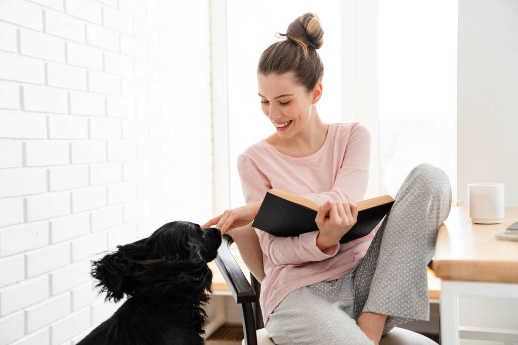 Усміхнена молода біла жінка в лаунж-одязі, сидячи з книгою на стільці вдома, грає зі своїм собакою
 - Фото, зображення