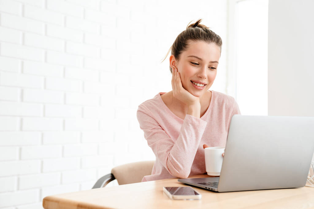 Lounge giysileri giyen mutlu beyaz kadın masada dizüstü bilgisayarla oturuyor, çalışıyor, video çekiyor. - Fotoğraf, Görsel