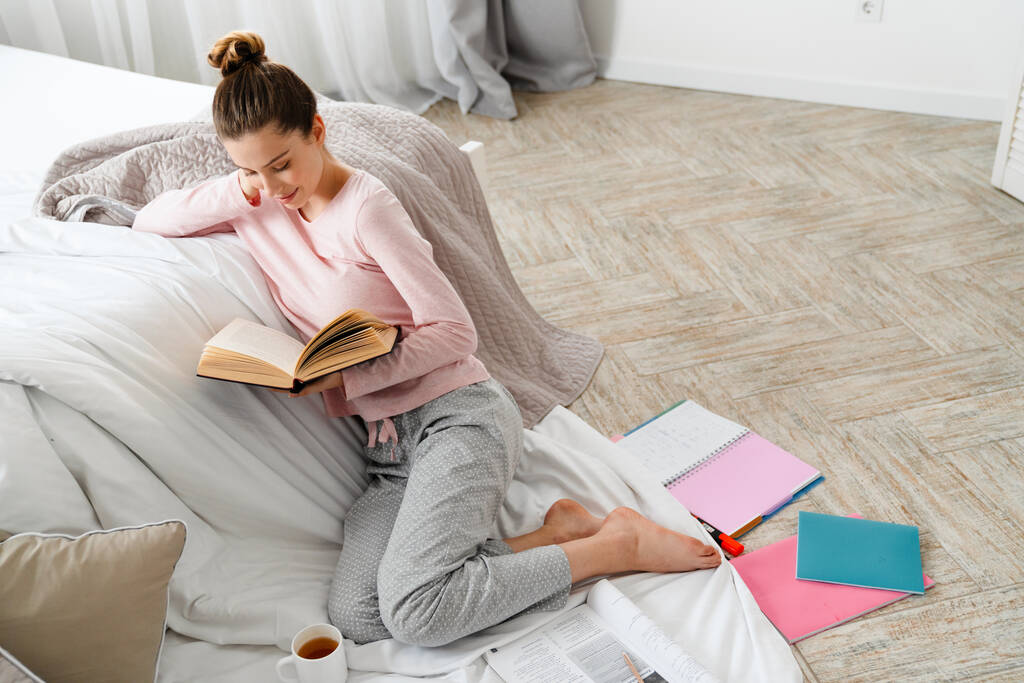 Joven mujer blanca leyendo libro estudiando sentada en el suelo de su dormitorio sobre una manta - Foto, Imagen