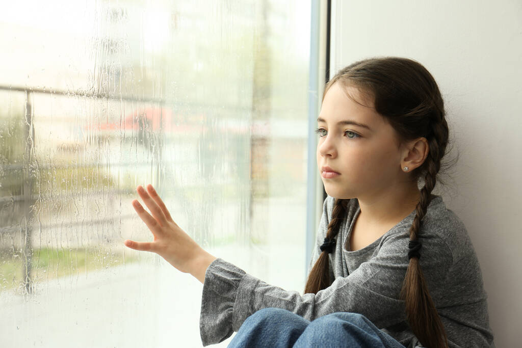 Ragazzina triste vicino alla finestra in casa in un giorno di pioggia - Foto, immagini