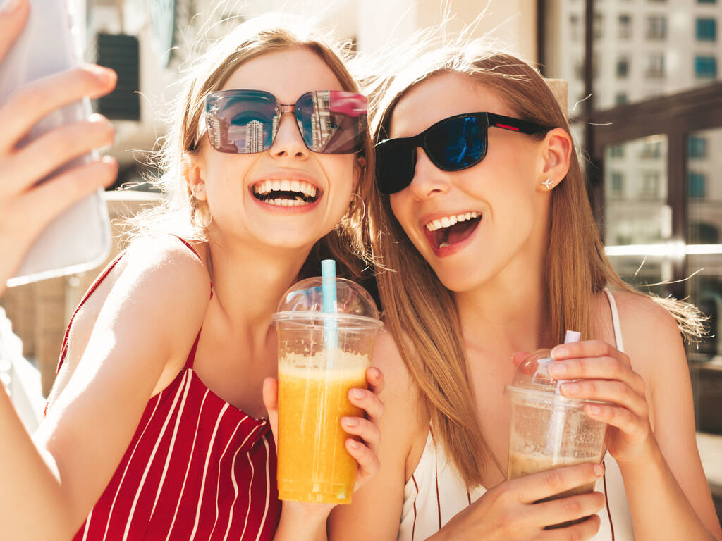 Twee jonge mooie glimlachende hippe teefjes in trendy zomerkleding. Zorgeloze vrouwen poseren buiten.Positieve modellen drinken verse cocktail smoothie drankje in plastic beker met stro. - Foto, afbeelding
