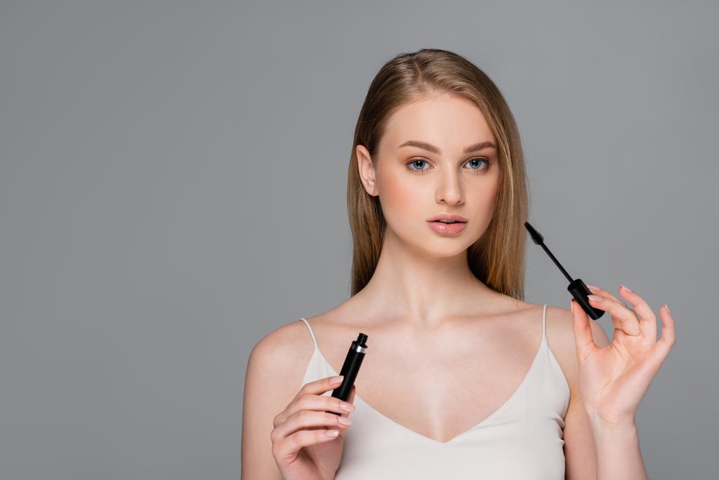 young woman holding tube with mascara and looking at camera isolated on grey - Photo, Image