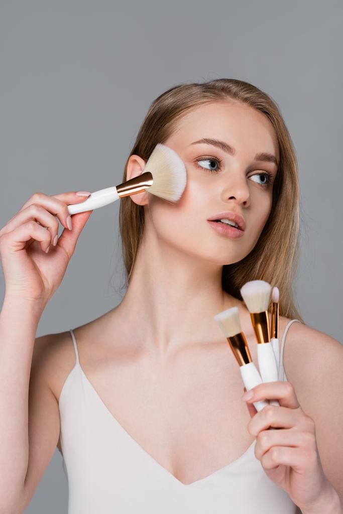 fiatal nő gazdaság készlet különböző kozmetikai kefék és alkalmazása arc por elszigetelt szürke - Fotó, kép