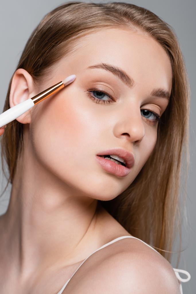 close up of young woman holding cosmetic brush and applying eye shadow isolated on grey - Photo, Image