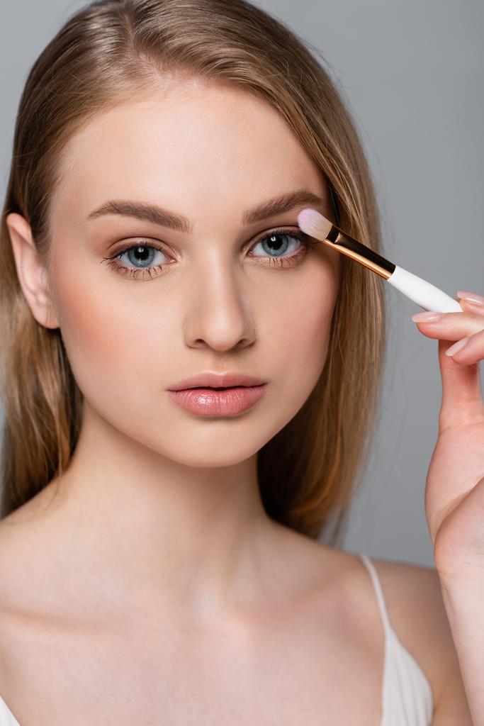 bonita mujer joven sosteniendo cepillo cosmético y aplicando sombra de ojos aislado en gris - Foto, Imagen