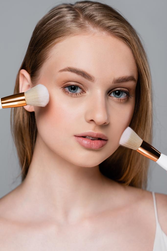 close up of young woman holding different cosmetic brushes and applying face powder isolated on grey - Photo, Image