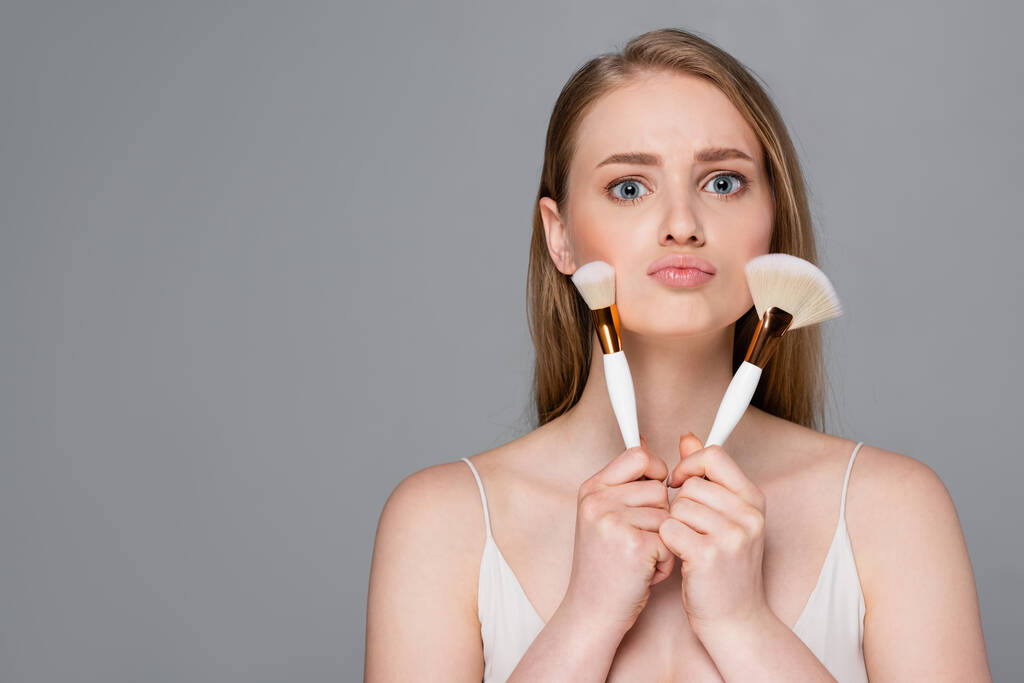 jovem segurando escovas cosméticas perto do rosto isolado em cinza - Foto, Imagem