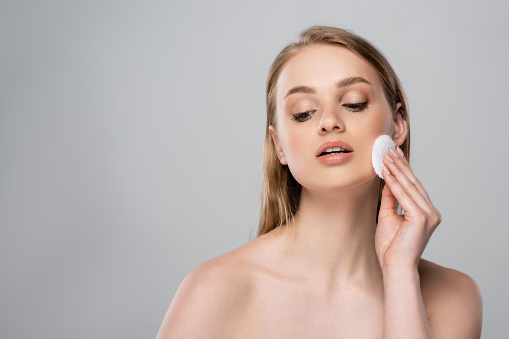 young woman with bare shoulders removing makeup with cotton pad isolated on gray - Photo, Image