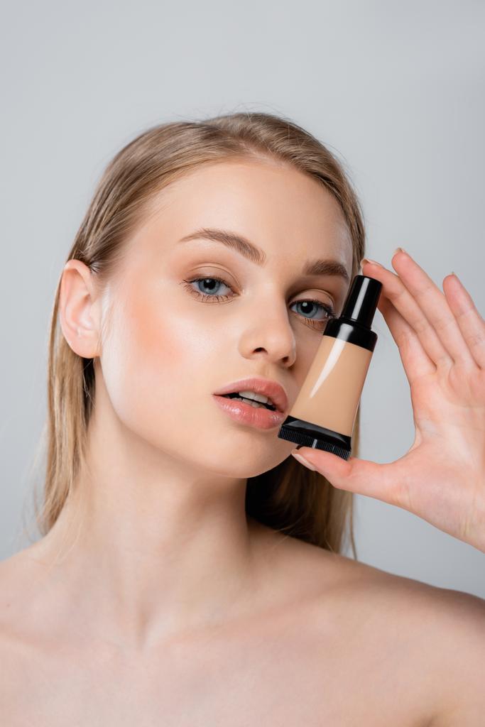 pretty woman with bare shoulders holding tube with makeup foundation isolated on grey  - Photo, Image