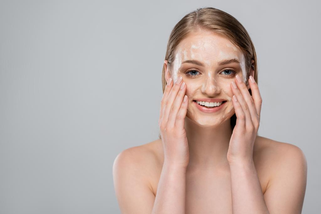 giovane donna positiva con occhi azzurri che applica schiuma detergente sul viso isolato su grigio - Foto, immagini