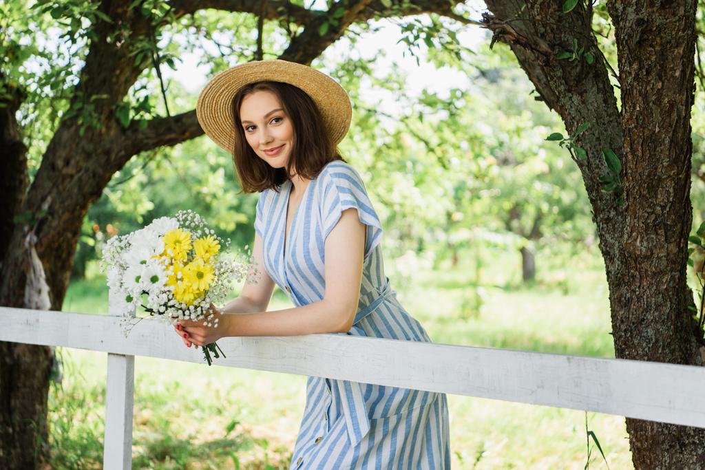 Mujer en sombrero de paja sosteniendo flores y sonriendo a la cámara cerca de la cerca  - Foto, imagen