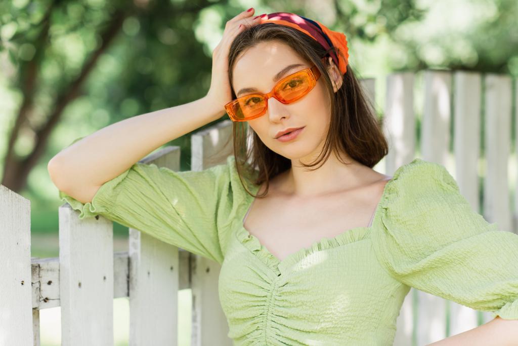 Hübsche Frau mit Kopftuch und Sonnenbrille steht am Zaun  - Foto, Bild