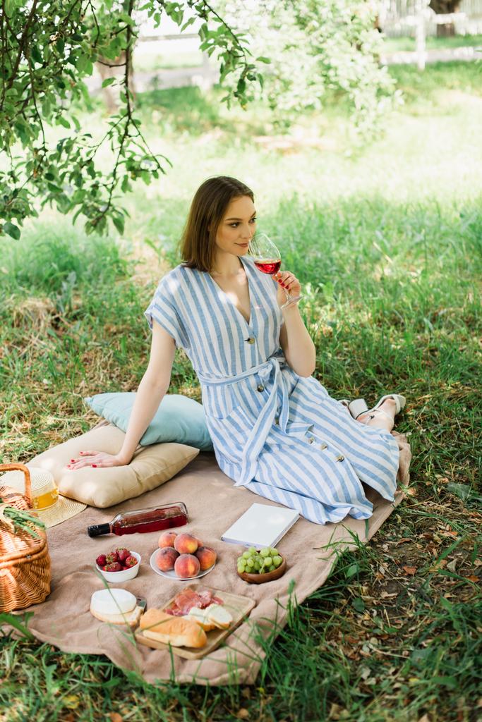 公園の毛布の上に食べ物、本、バスケットの近くにワインのガラスを保持する若い女性  - 写真・画像