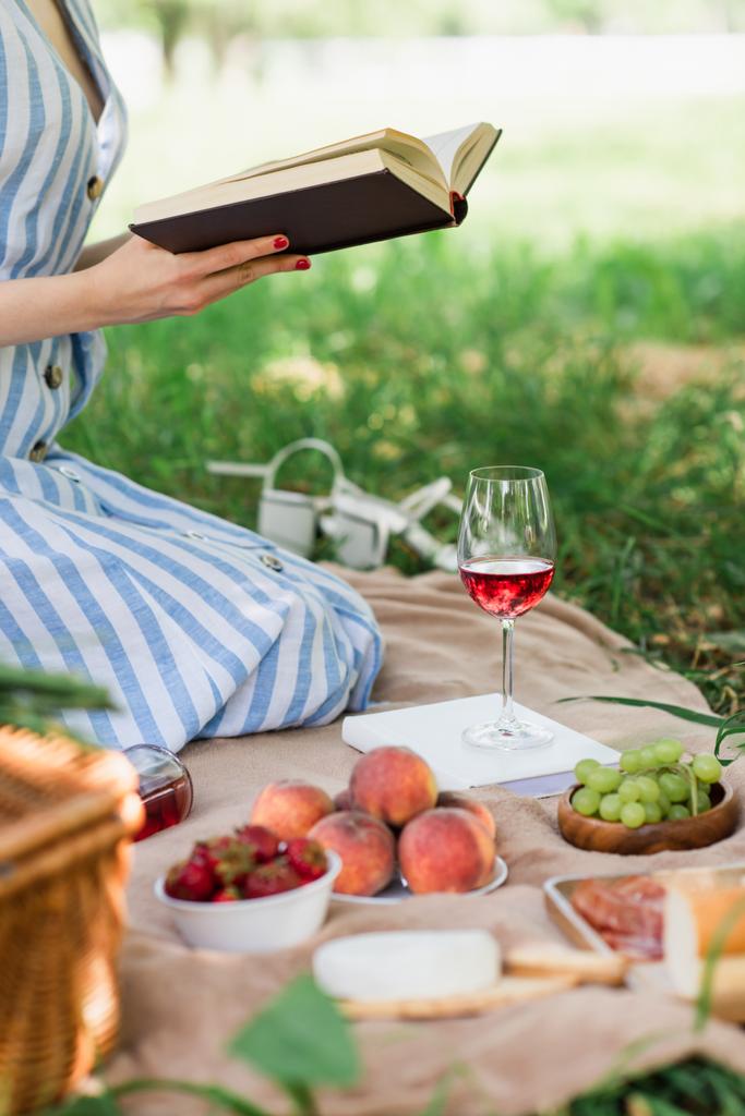 Widok kobiety czytającej książki w pobliżu żywności i wina w parku  - Zdjęcie, obraz
