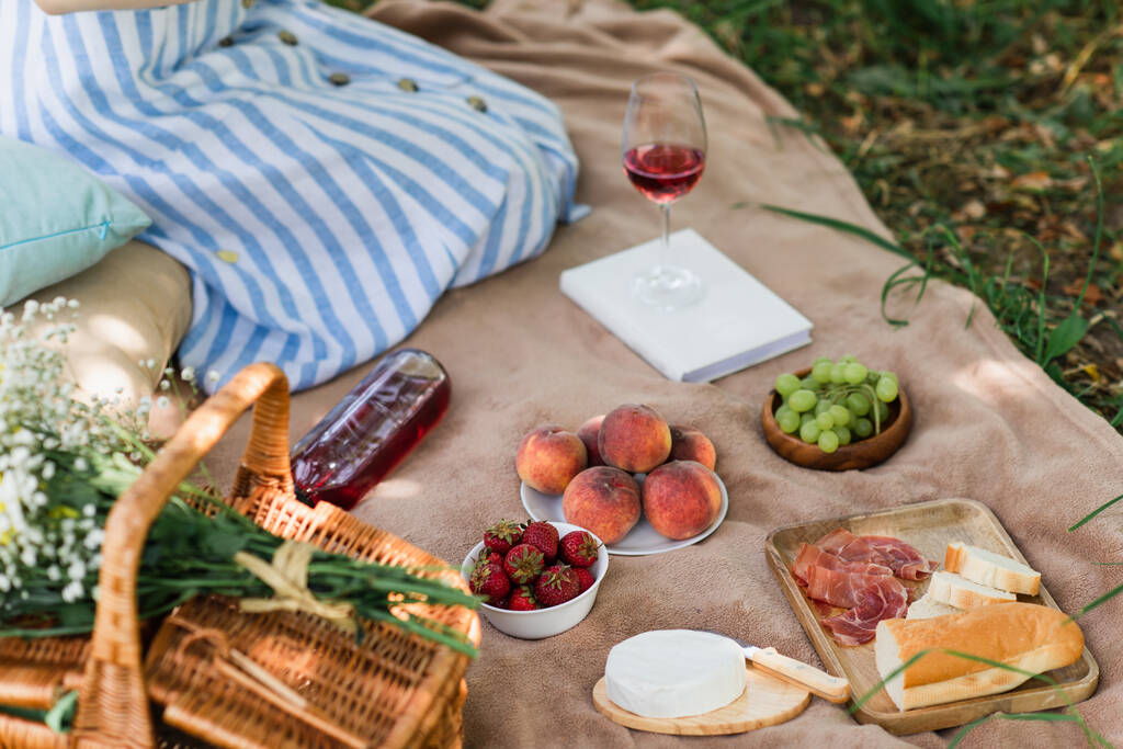Καλλιεργημένη θέα νόστιμο φαγητό και κρασί κοντά στο καλάθι και γυναίκα σε κουβέρτα στο πάρκο  - Φωτογραφία, εικόνα