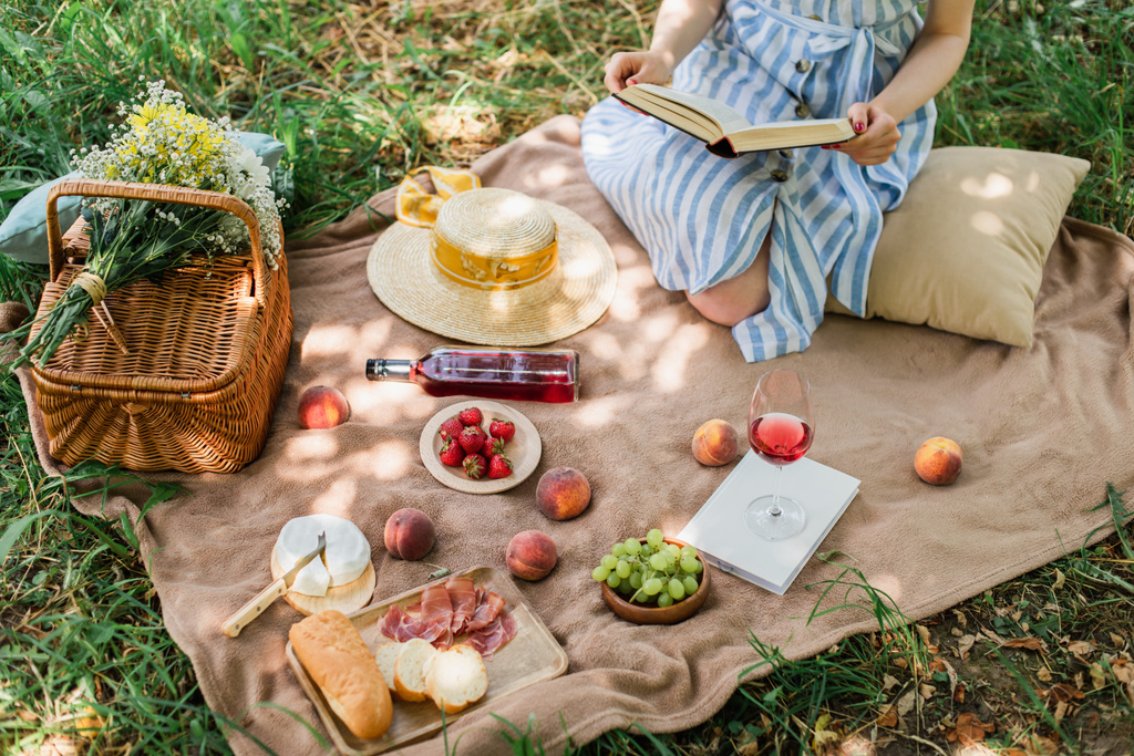 Widok na młodą kobietę trzymającą książkę podczas pikniku z winem i jedzeniem w parku  - Zdjęcie, obraz