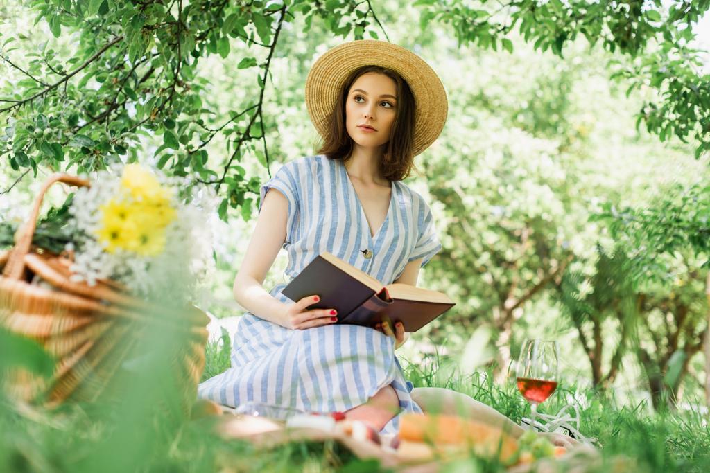 Donna in cappello di paglia che tiene libro vicino al vino e cesto sfocato nel parco  - Foto, immagini