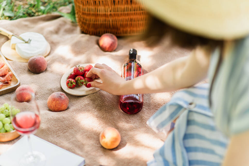 Widok kobiety biorącej truskawki w pobliżu wina i owoców na kocu w parku  - Zdjęcie, obraz