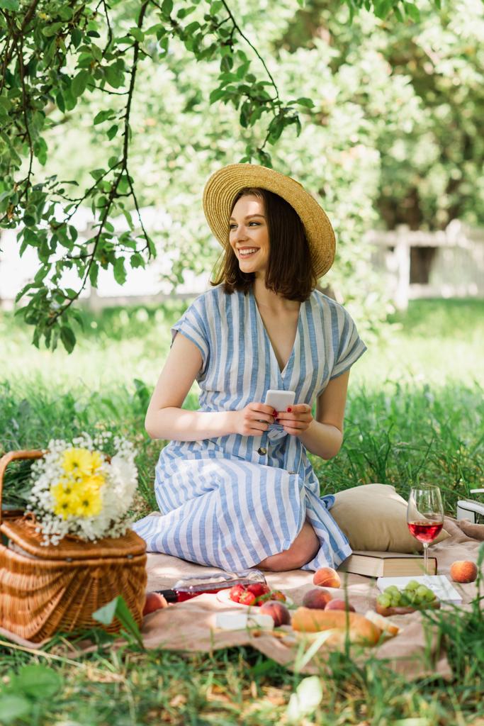 Smiling woman holding smartphone near food and wine on blanket in park  - Photo, Image