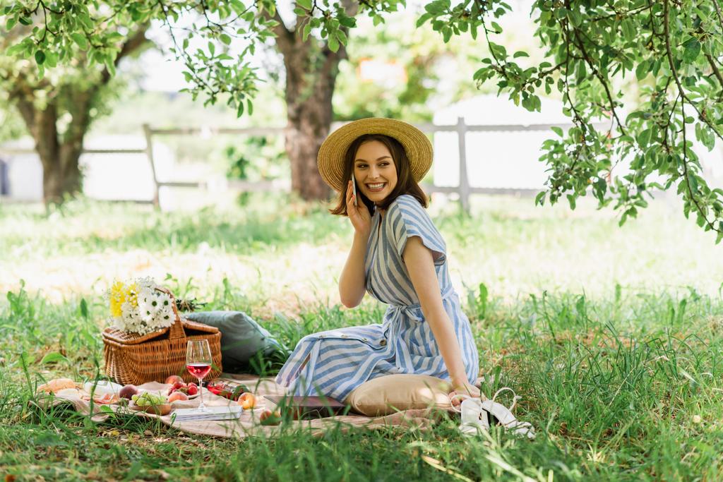 公園でワイン、食べ物や花の近くに携帯電話で話す太陽の帽子の女性  - 写真・画像