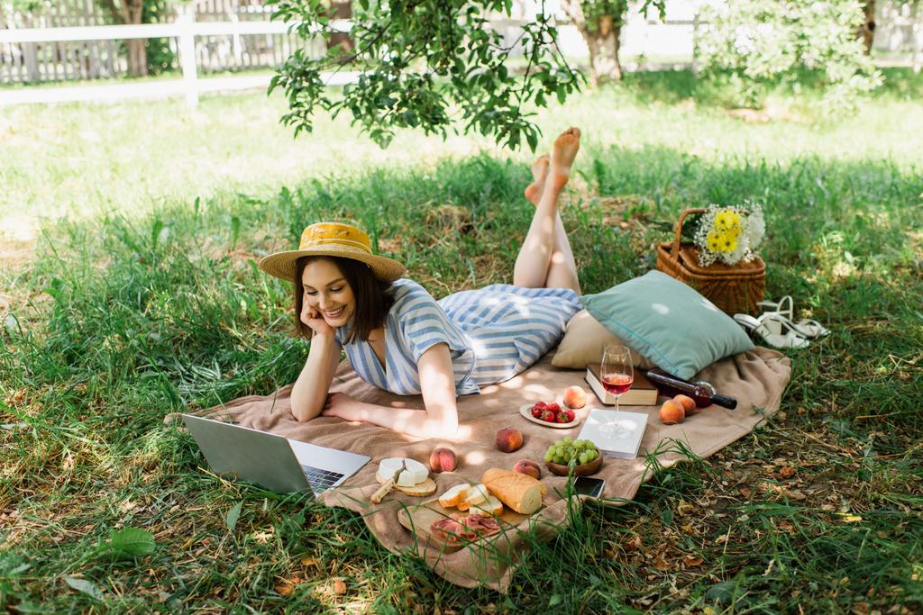 Uśmiechnięta kobieta patrząca na laptopa w pobliżu książek, wina i jedzenia na kocu piknikowym  - Zdjęcie, obraz