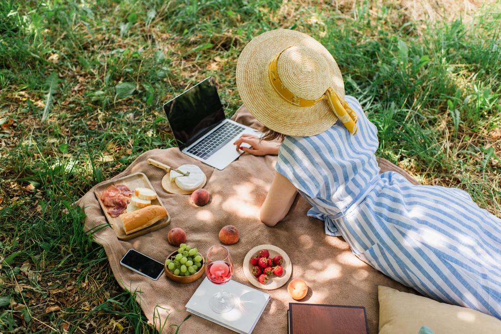 Vista superior da mulher usando laptop perto de smartphone, comida e vinho no cobertor no parque  - Foto, Imagem