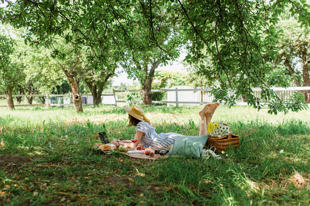 Босоногая женщина лежит рядом с ноутбуком, еда и вино на одеяле в парке  - Фото, изображение