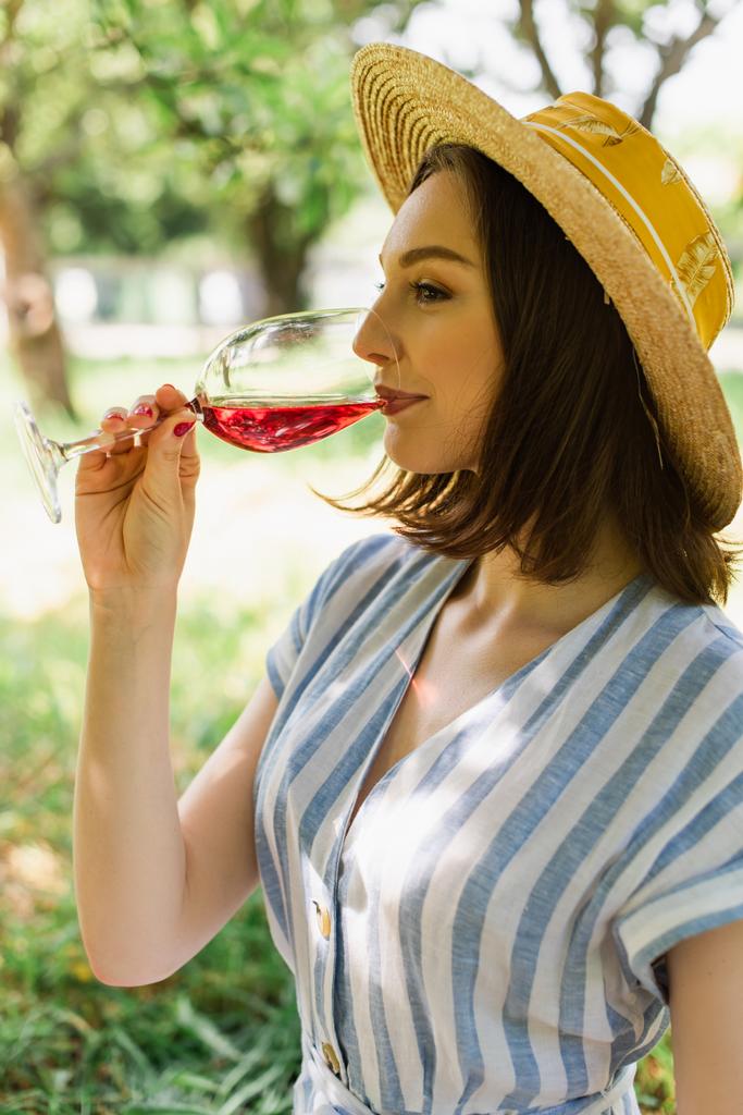 Πλευρική άποψη της γυναίκας στον ήλιο καπέλο πίνοντας κρασί στο πάρκο  - Φωτογραφία, εικόνα