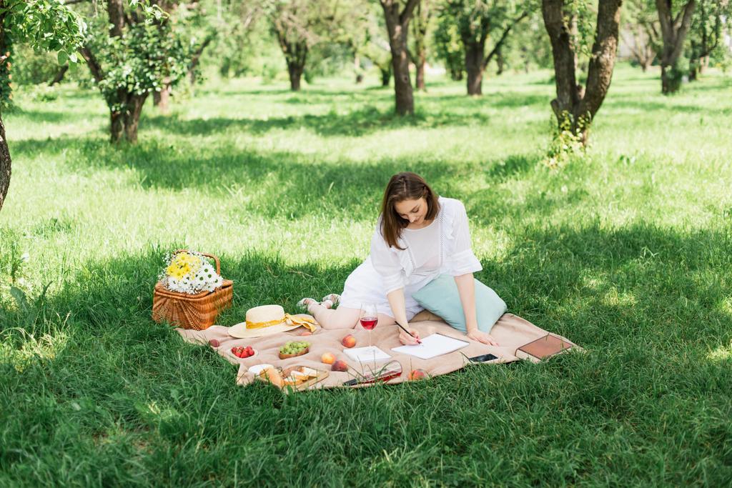 公園で毛布の上に食べ物やワインの近くに描画若い女性  - 写真・画像