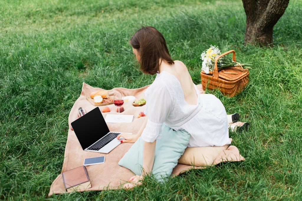 Mujer joven usando el ordenador portátil cerca de teléfono inteligente, libros y comida en la manta de picnic  - Foto, imagen