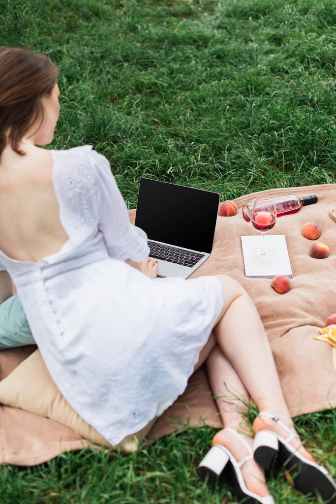 Mujer borrosa usando portátil con pantalla en blanco cerca del vino y frutas en la hierba en el parque  - Foto, imagen