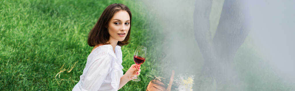 Młoda kobieta trzyma kieliszek wina w pobliżu kosza i kwiaty na trawie w parku, sztandar  - Zdjęcie, obraz