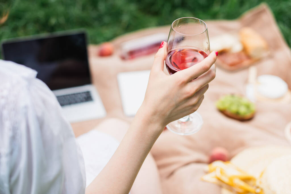 Cropped view of woman holding glass of wine on blurred blanket in park  - Photo, Image
