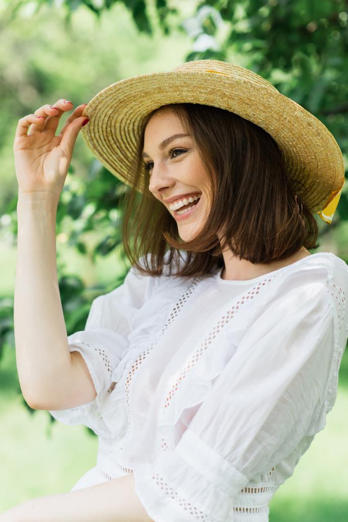 Donna sorridente che tiene il cappello di paglia e distoglie lo sguardo nel parco  - Foto, immagini