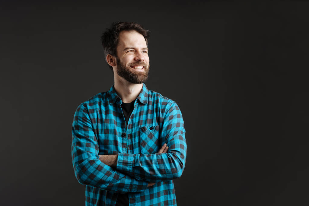Bearded brunette man in shirt smiling and looking aside isolated over black background - Photo, Image